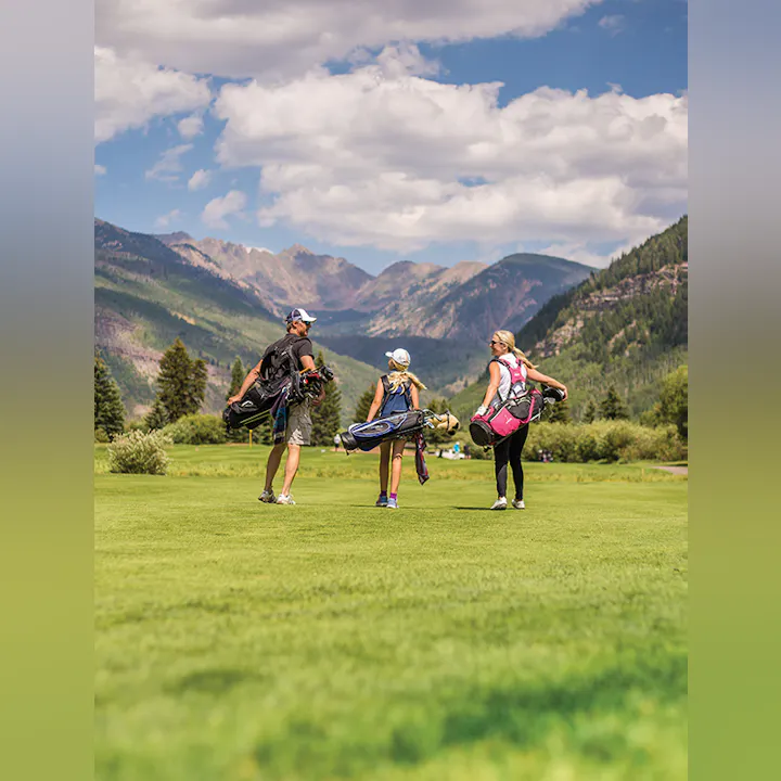 Three golfers with bags on a lush course, mountains in the background.