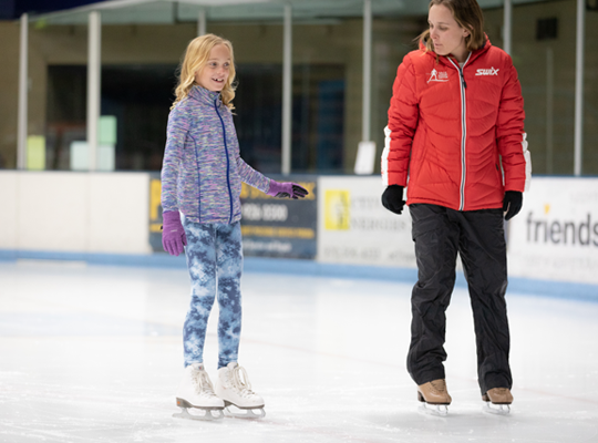 Two people ice skating, one appears to be teaching or assisting the other.