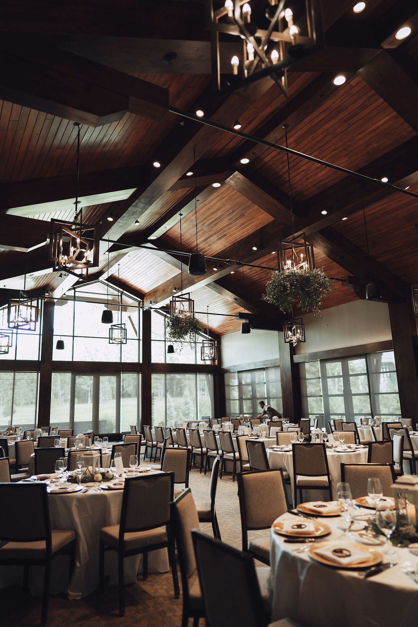Elegant dining hall with wooden beams, chandeliers, set tables, and large windows.