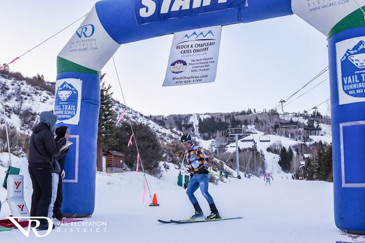 A skier starting a race through a blue arch in a snowy mountain setting, with onlookers and a visible chairlift.