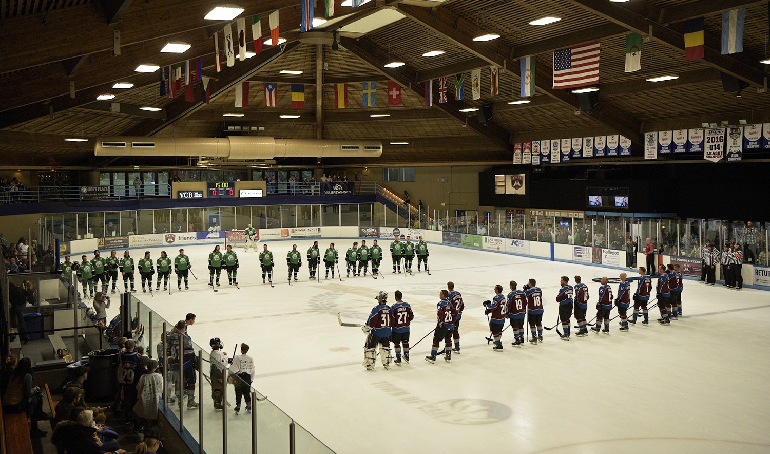 Two ice hockey teams lined up on the rink, ready for a game, with spectators and flags visible in an indoor arena.