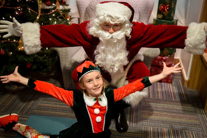 Santa and an elf pose joyfully in a festive setting with a Christmas tree and gifts.
