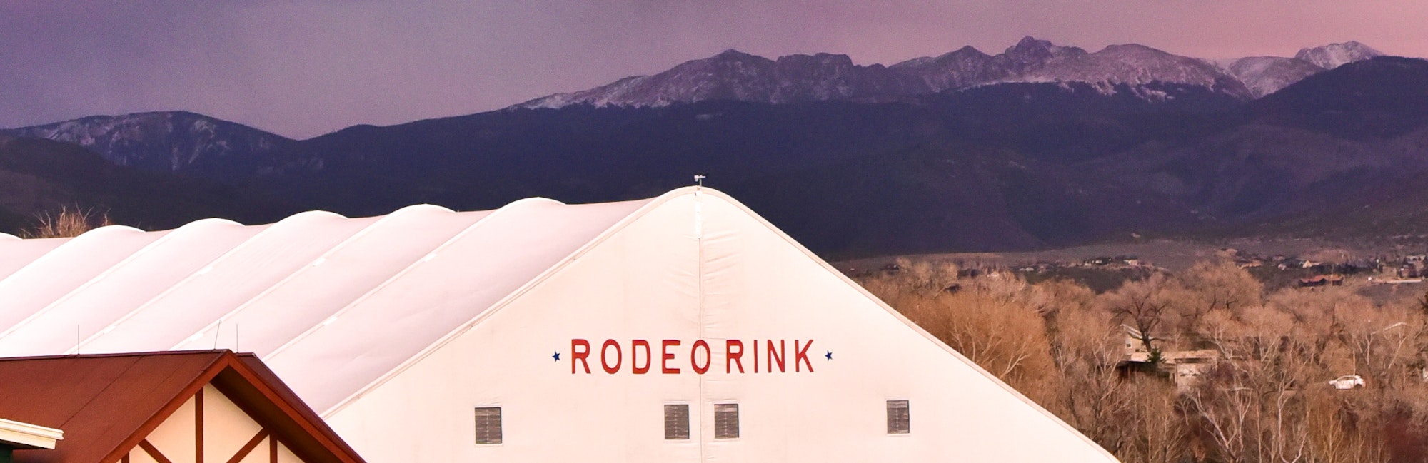 The image features a large white building labeled "RODEO RINK" with mountains in the background.