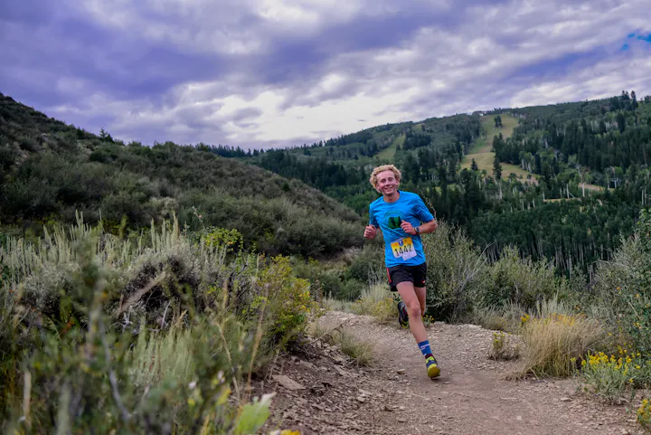 A runner with a bib number on a mountain trail, surrounded by greenery under a cloudy sky.