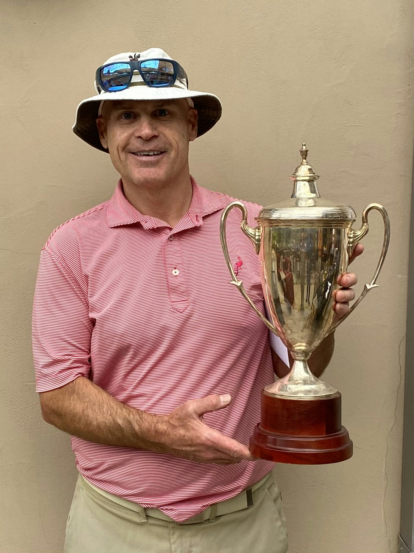A person smiling, holding a trophy, wearing a hat with sunglasses atop, and a striped pink polo shirt.