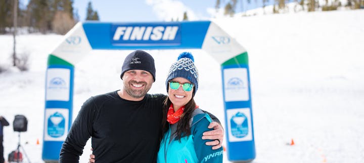 Two people smiling in front of a "FINISH" arch at a snowy location.