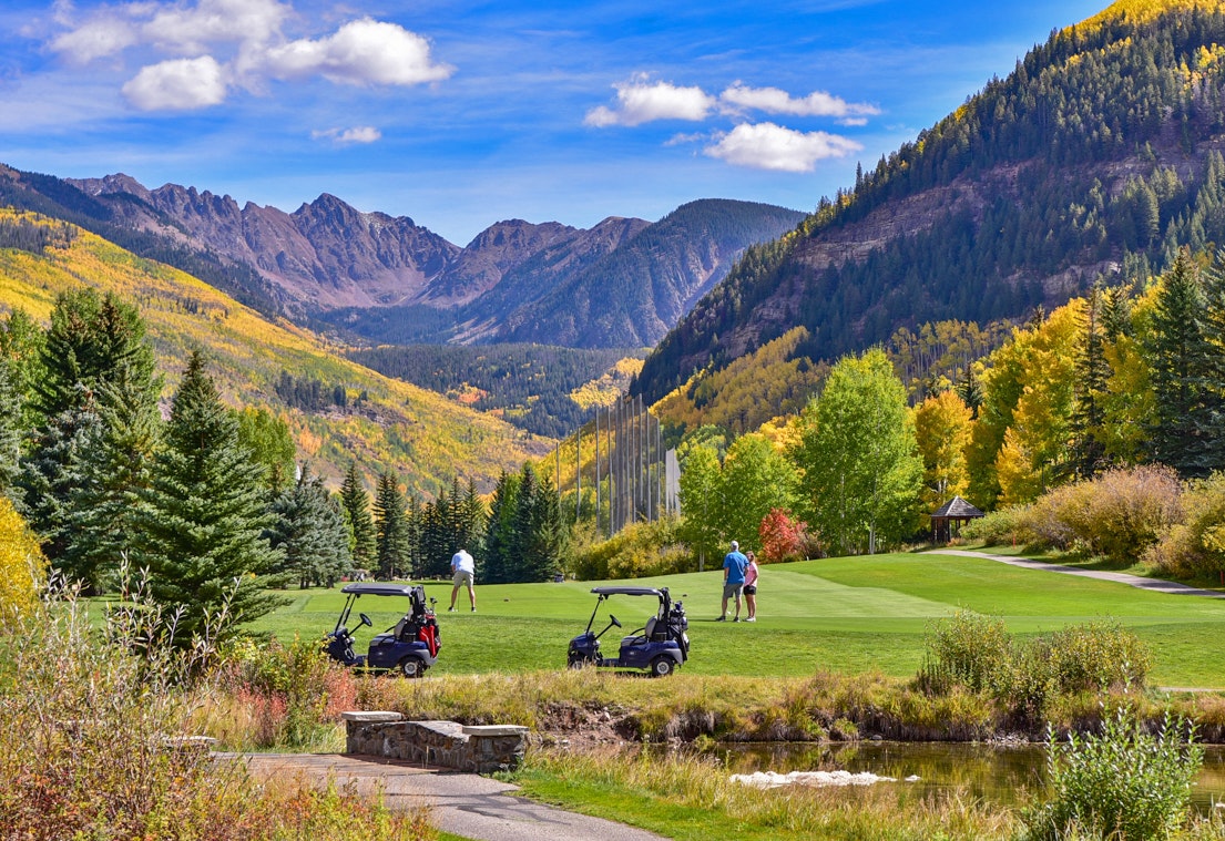 Golfers on a green with golf carts, surrounded by colorful autumn trees and mountains under a blue sky.