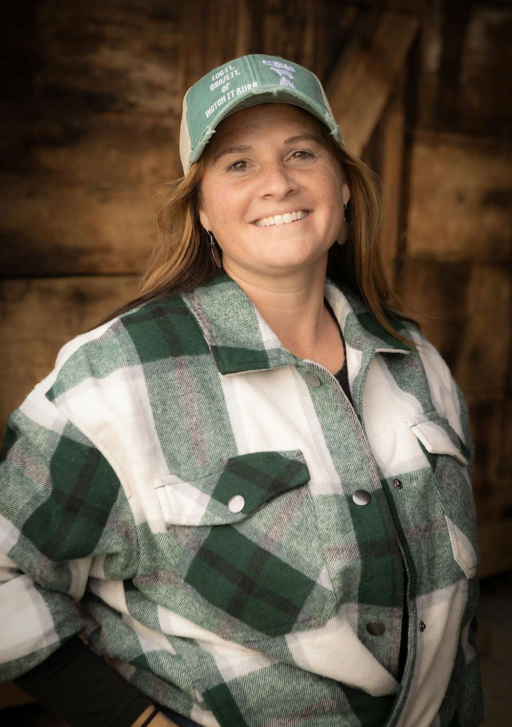 A smiling person in a green cap and plaid shirt standing in front of a wooden background.