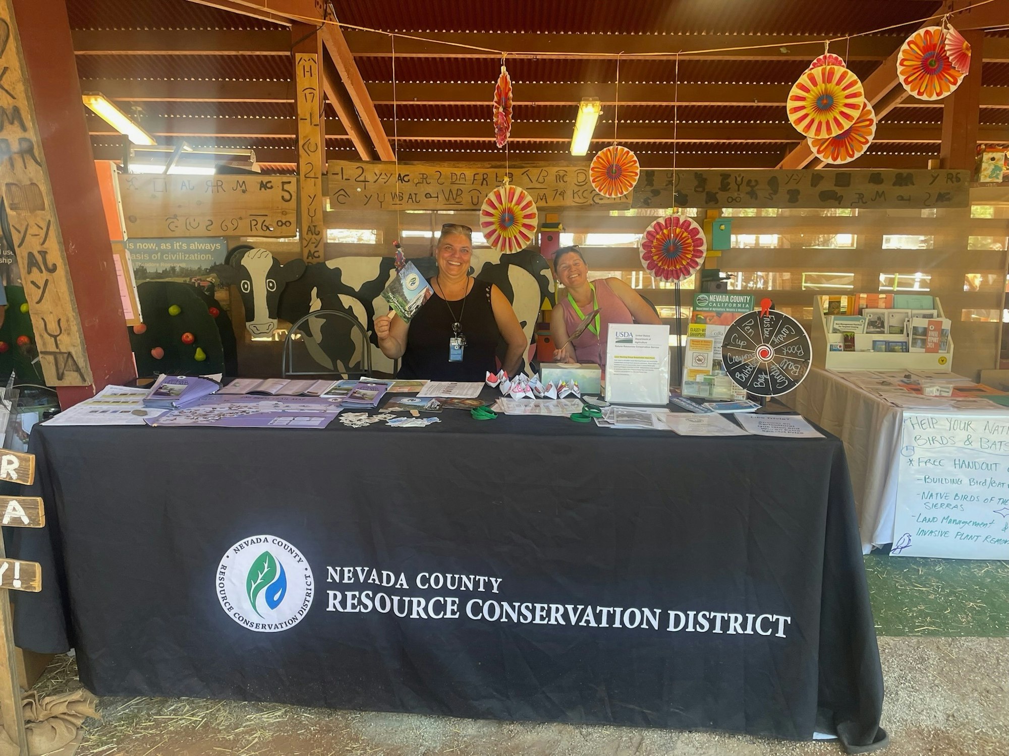 The image shows a booth for Nevada County Resource Conservation District with two smiling women and various informational materials.