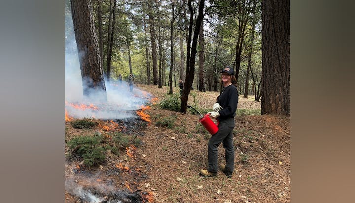 A person with a red canister near a controlled fire in a forest, smiling at the camera.