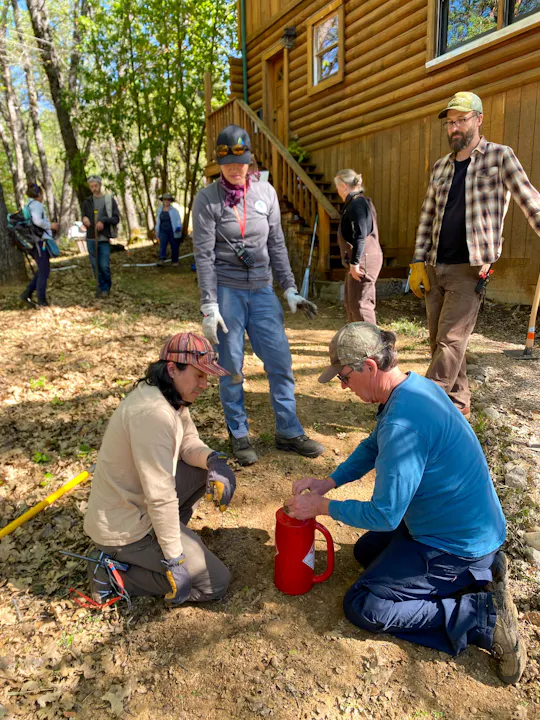A group of people is outdoors, preparing the area for a prescribed burn, near a log cabin.