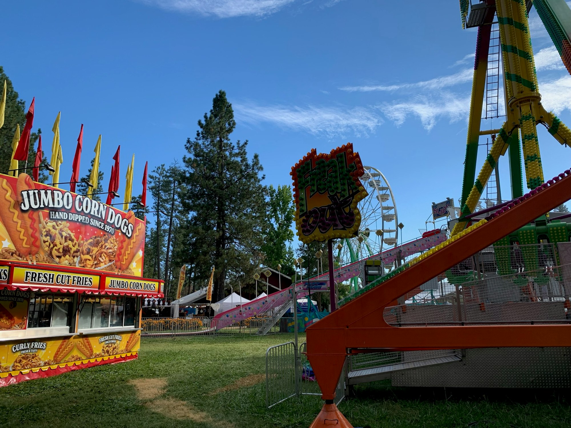 The image shows a fair scene with food stalls, rides, and colorful flags under a blue sky. Main attractions include corn dogs and a giant wheel.