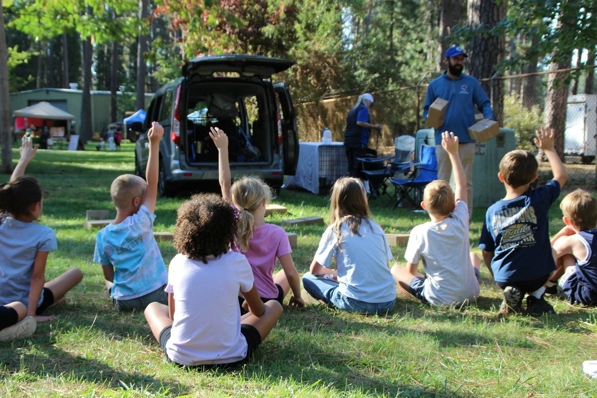 A group of children sitting in a grassy area, raising hands, while an adult presents nearby, likely involved in an outdoor activity.