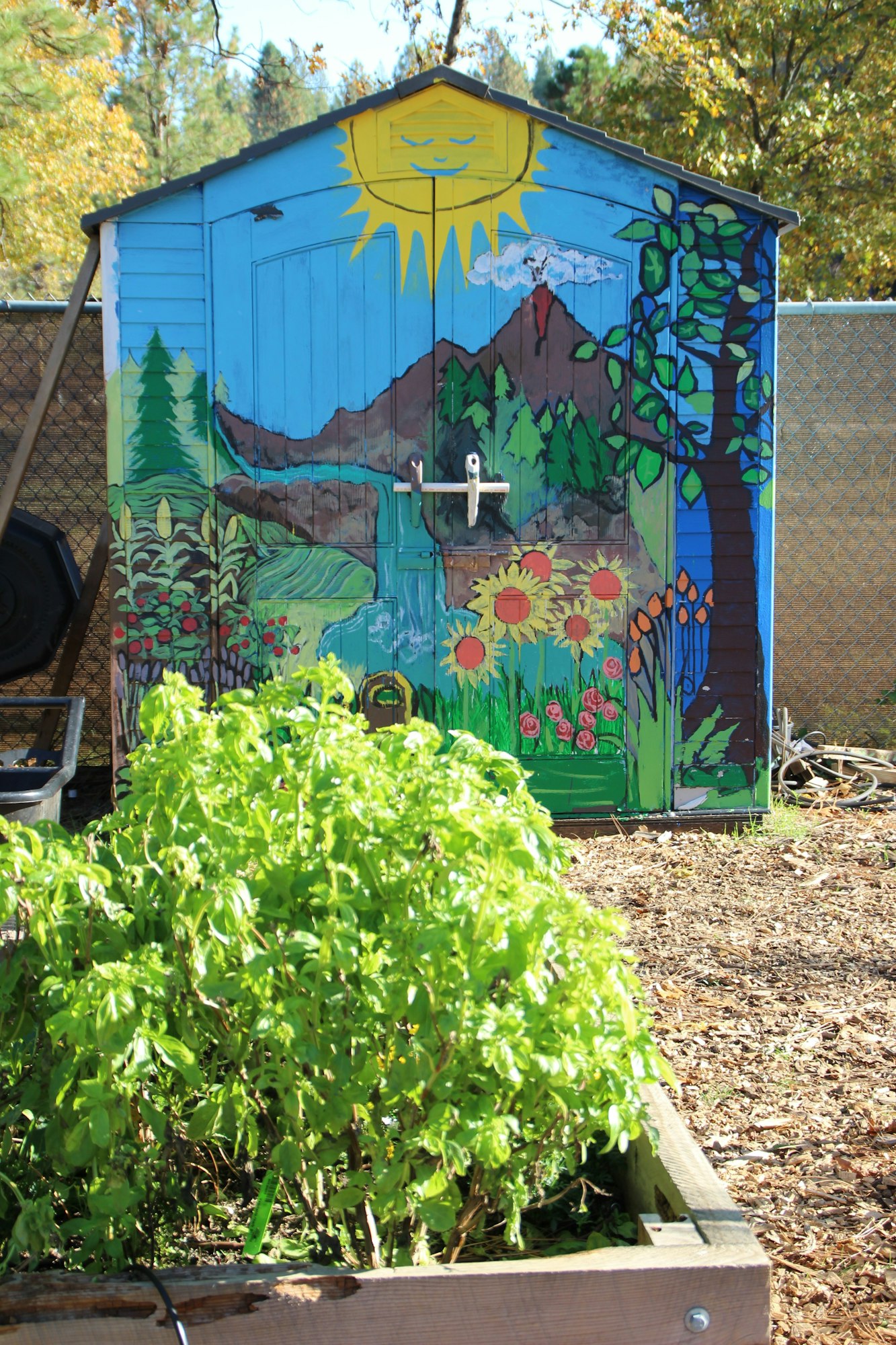 A painted garden shed features a sunny landscape with trees, flowers, and a mountain, surrounded by lush green plants.