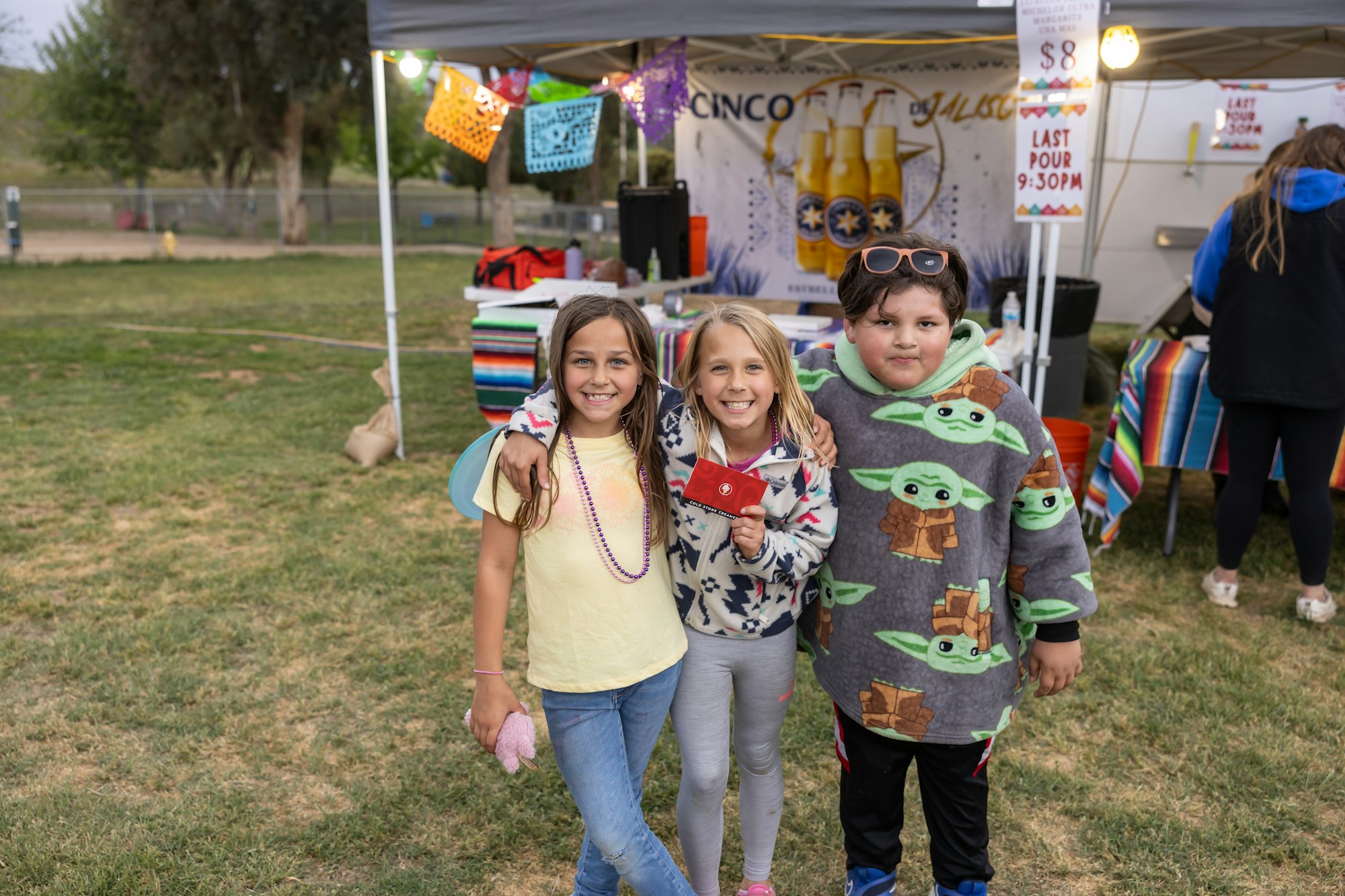 Three children smiling, standing in front of a tent with colorful decorations and a banner in a grassy area.