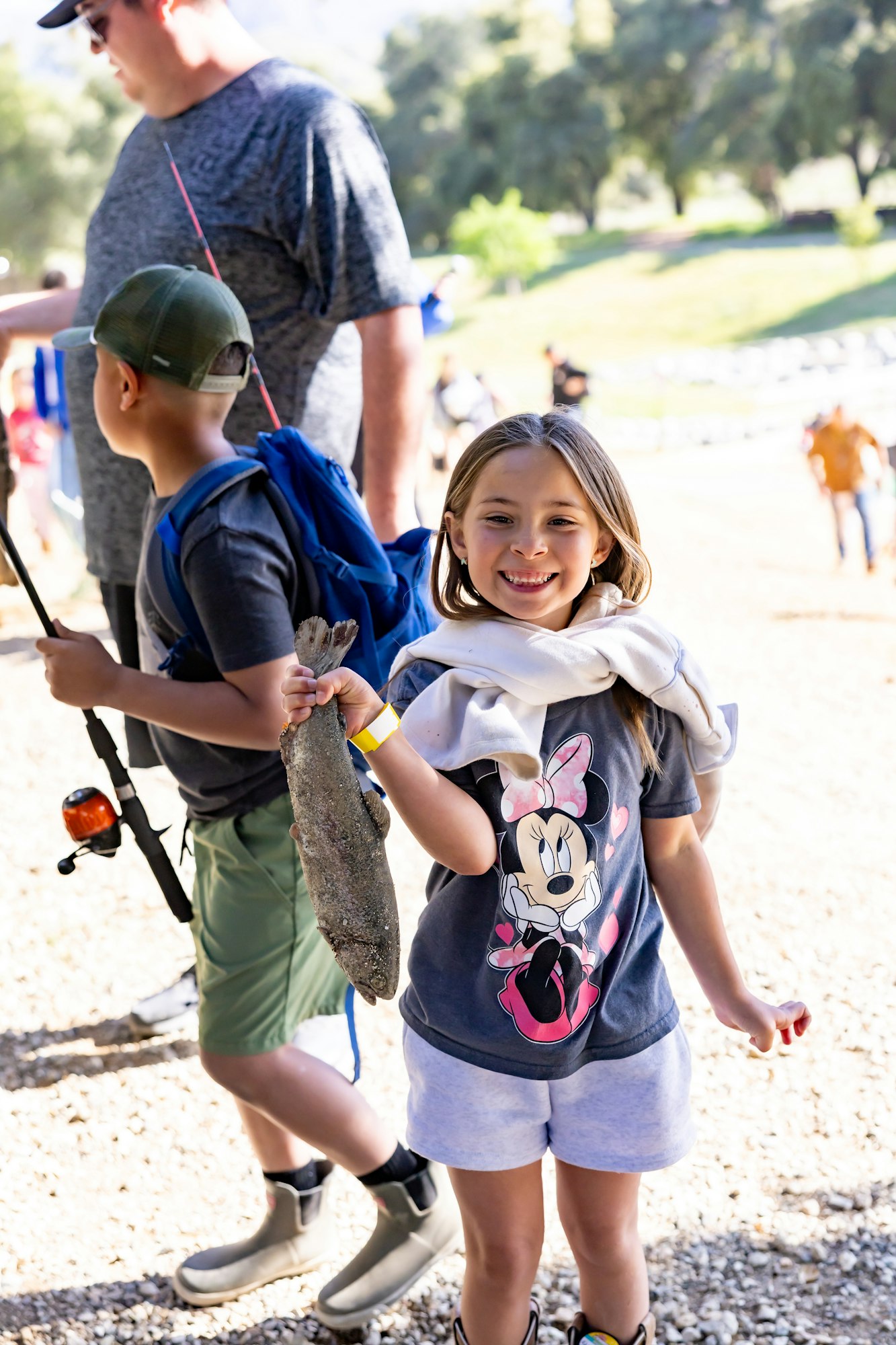 Smiling girl holds a fish; boy and man with fishing gear in background, outdoors.