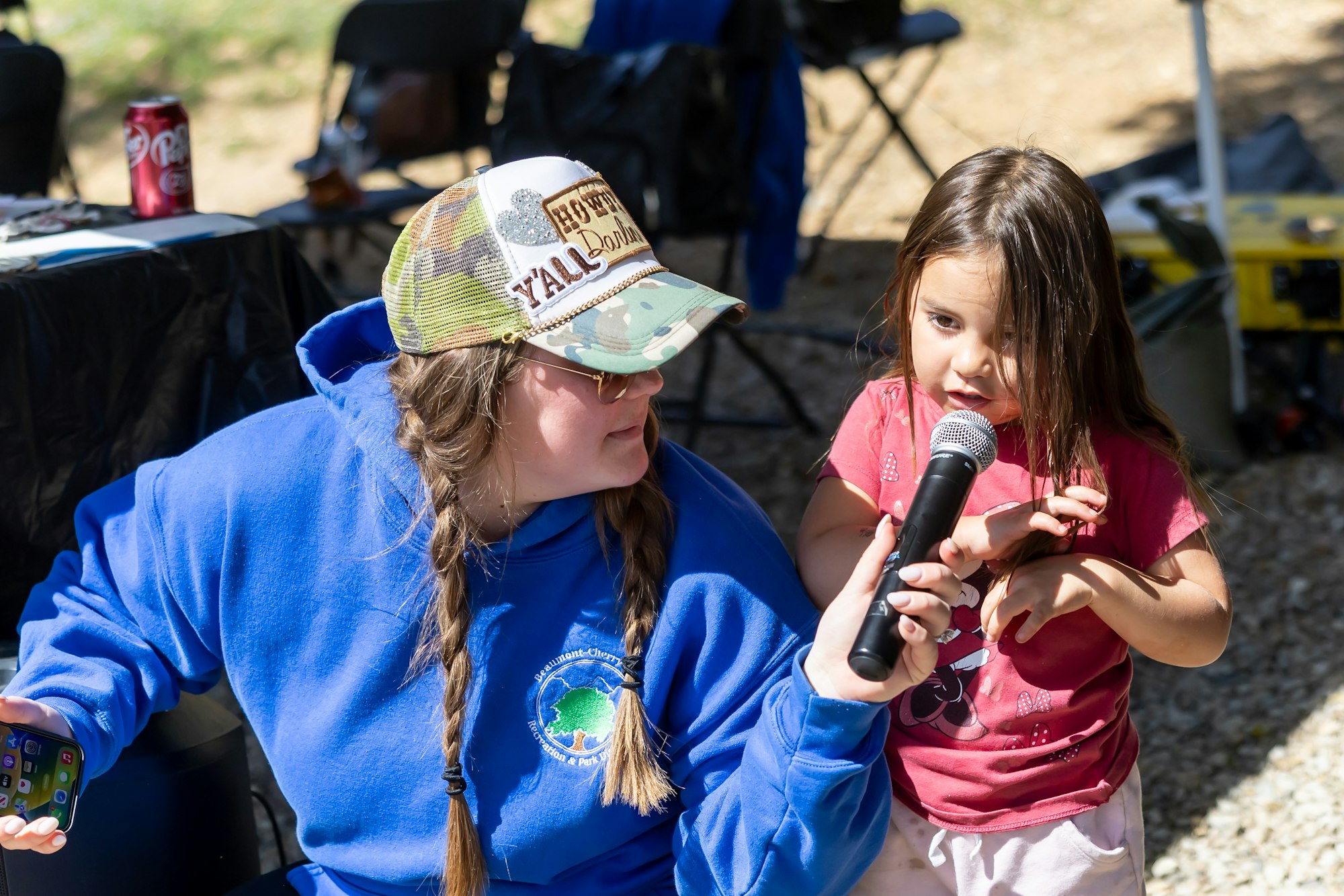 A person in a blue hoodie helps a child with a microphone outdoors.