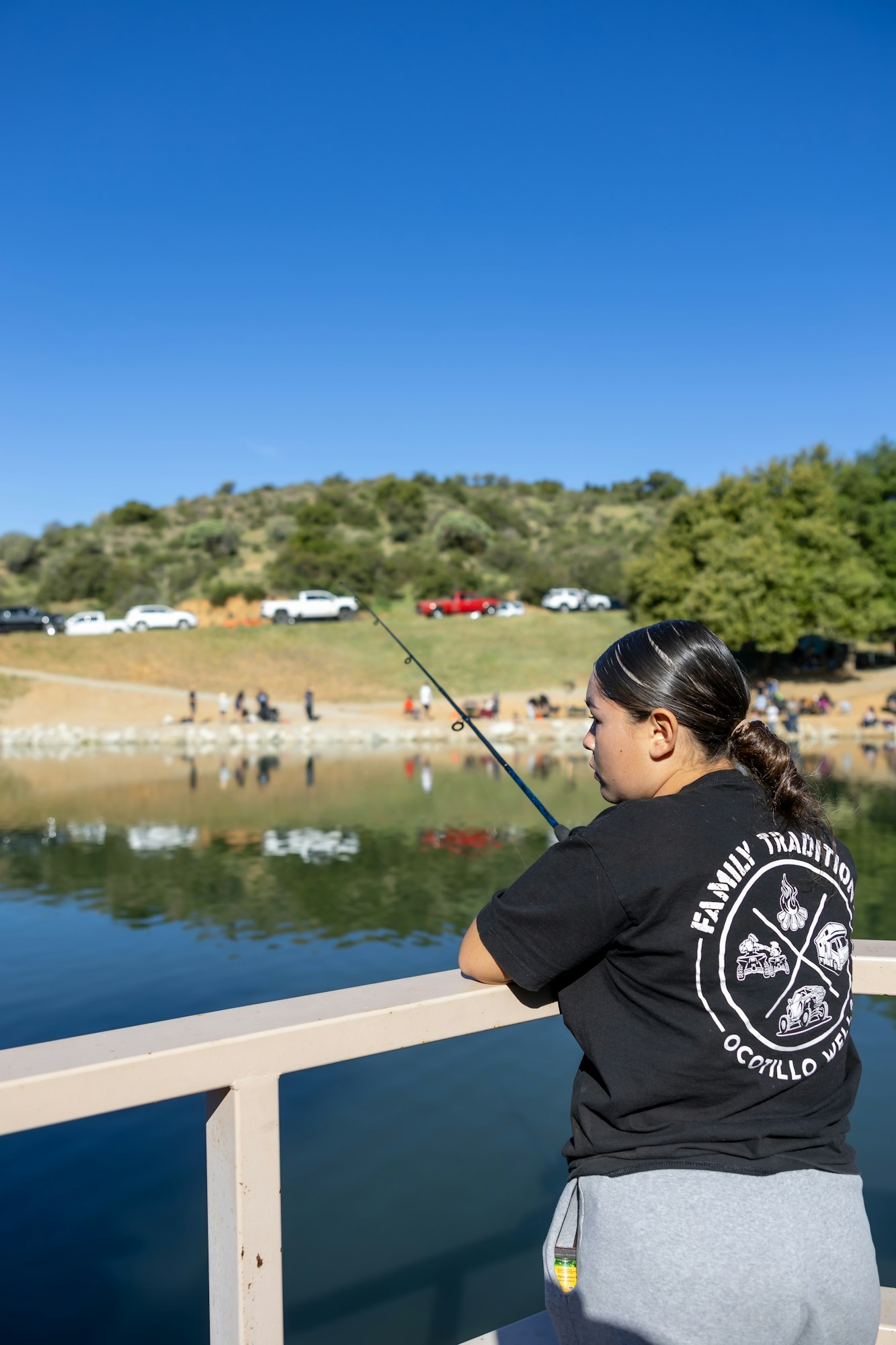 Person fishing on a railing by a lake, with people and cars in the background.