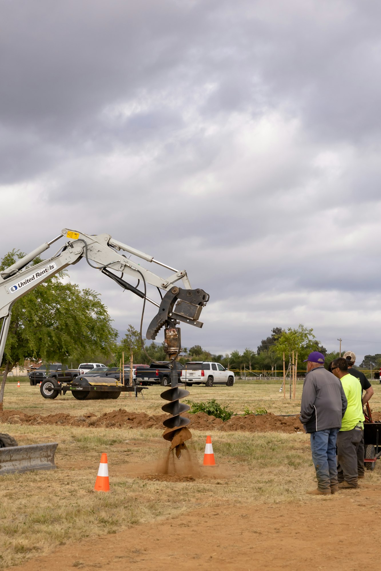 Construction site with excavator auger drilling into the ground, workers observing, and traffic cones marking the area.