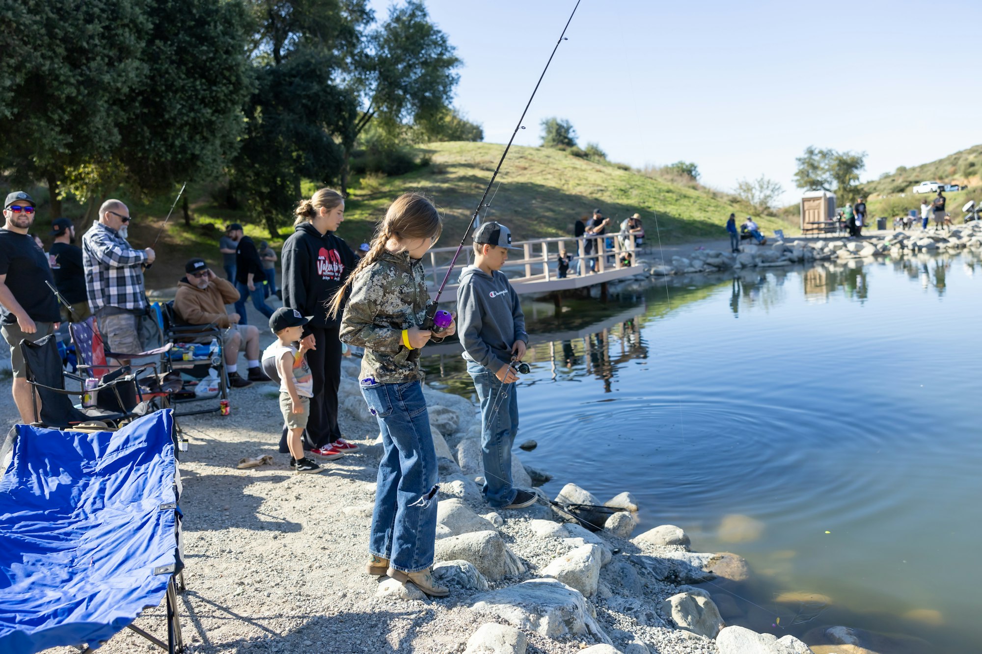 People fishing by a lakeside, with many individuals gathered, some with fishing rods, enjoying a calm outdoor setting.