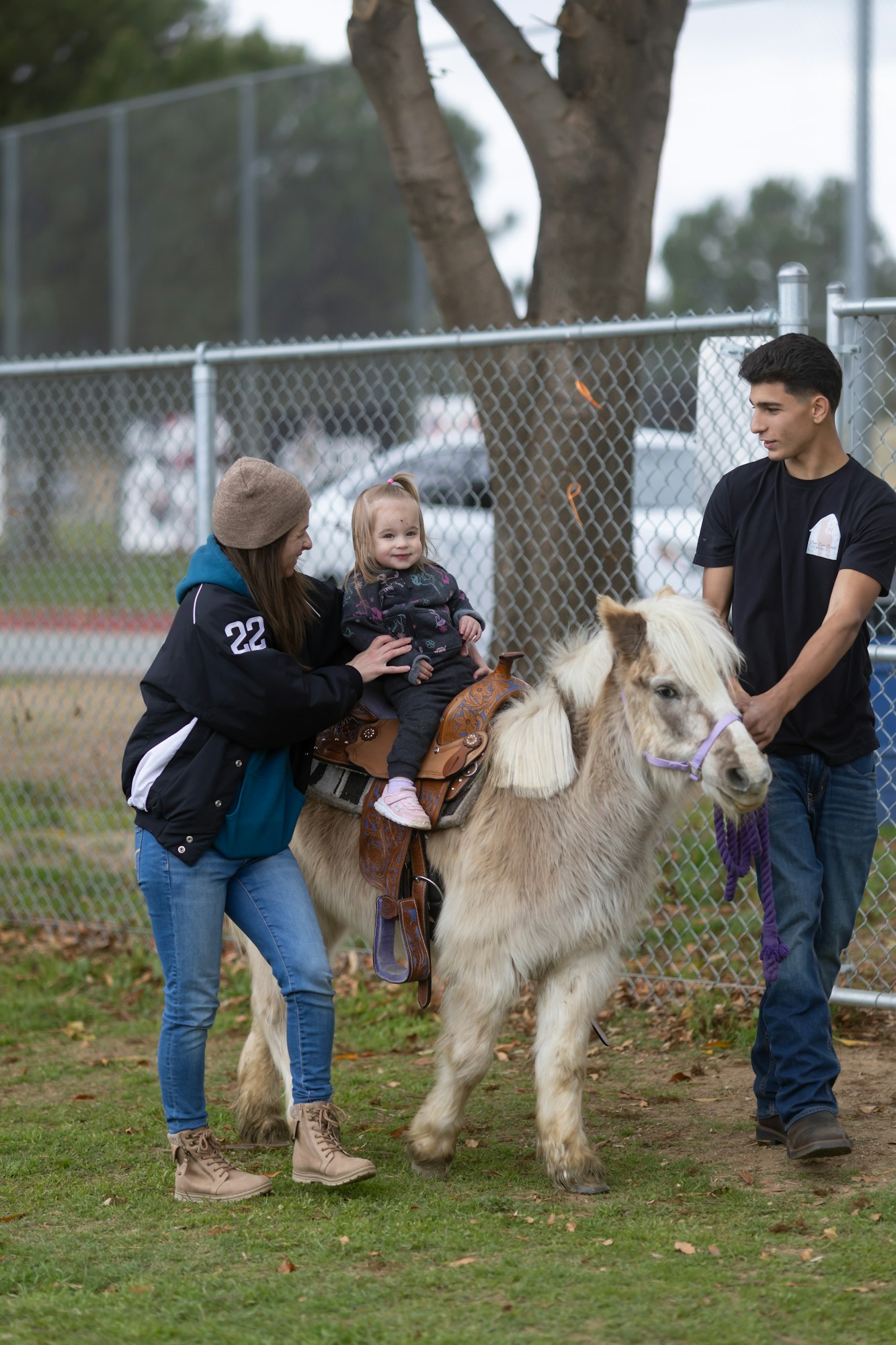 A child is seated on a pony, guided by an adult, with another person holding the pony's lead.