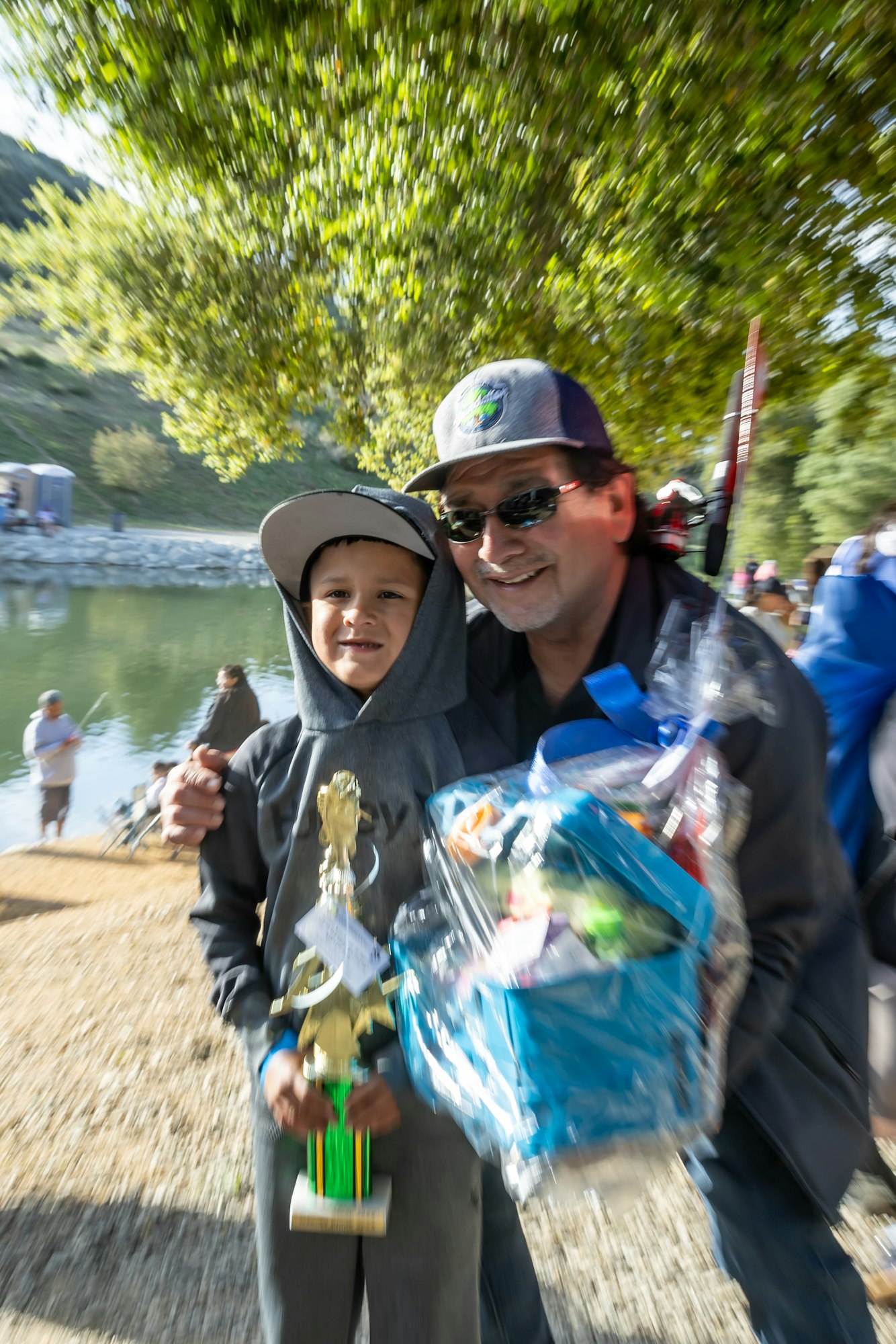 A person and a child near a lake, holding a trophy and gift basket, smiling.