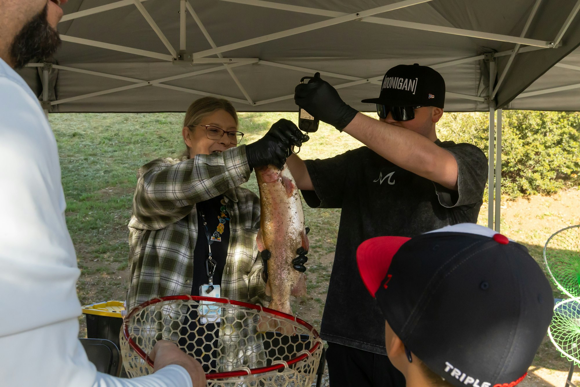 A person is holding a fish with tongs while someone else takes a photo under a canopy. A child is watching nearby.