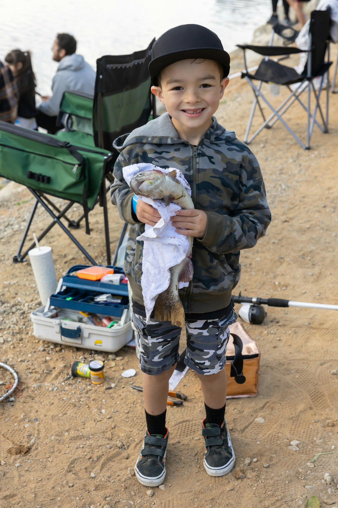 A smiling child holds a fish wrapped in a towel, standing on a sandy area with fishing gear and foldable chairs nearby.