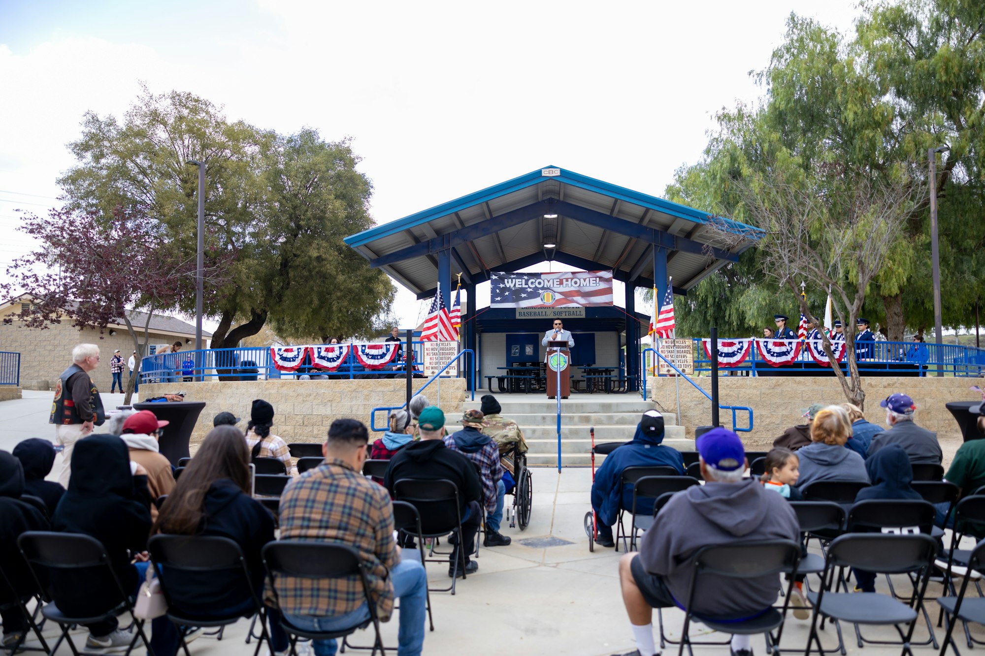 Outdoor event with people seated facing a speaker on a decorated stage with "WELCOME HOME!" and U.S. flags.