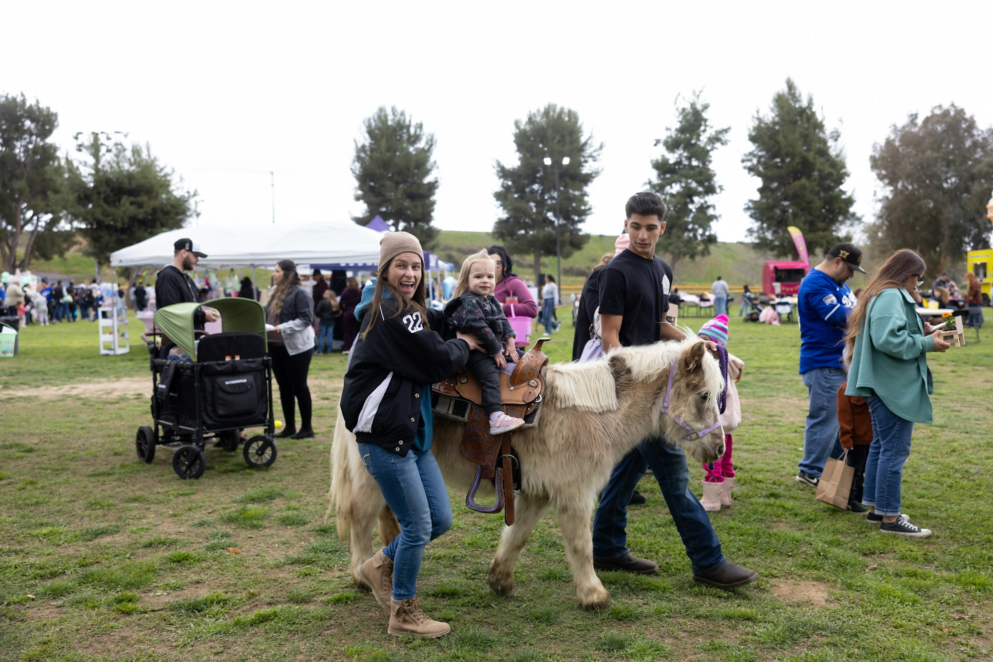 A child is riding a pony with an adult guiding, surrounded by people at an outdoor event with tents and strollers.