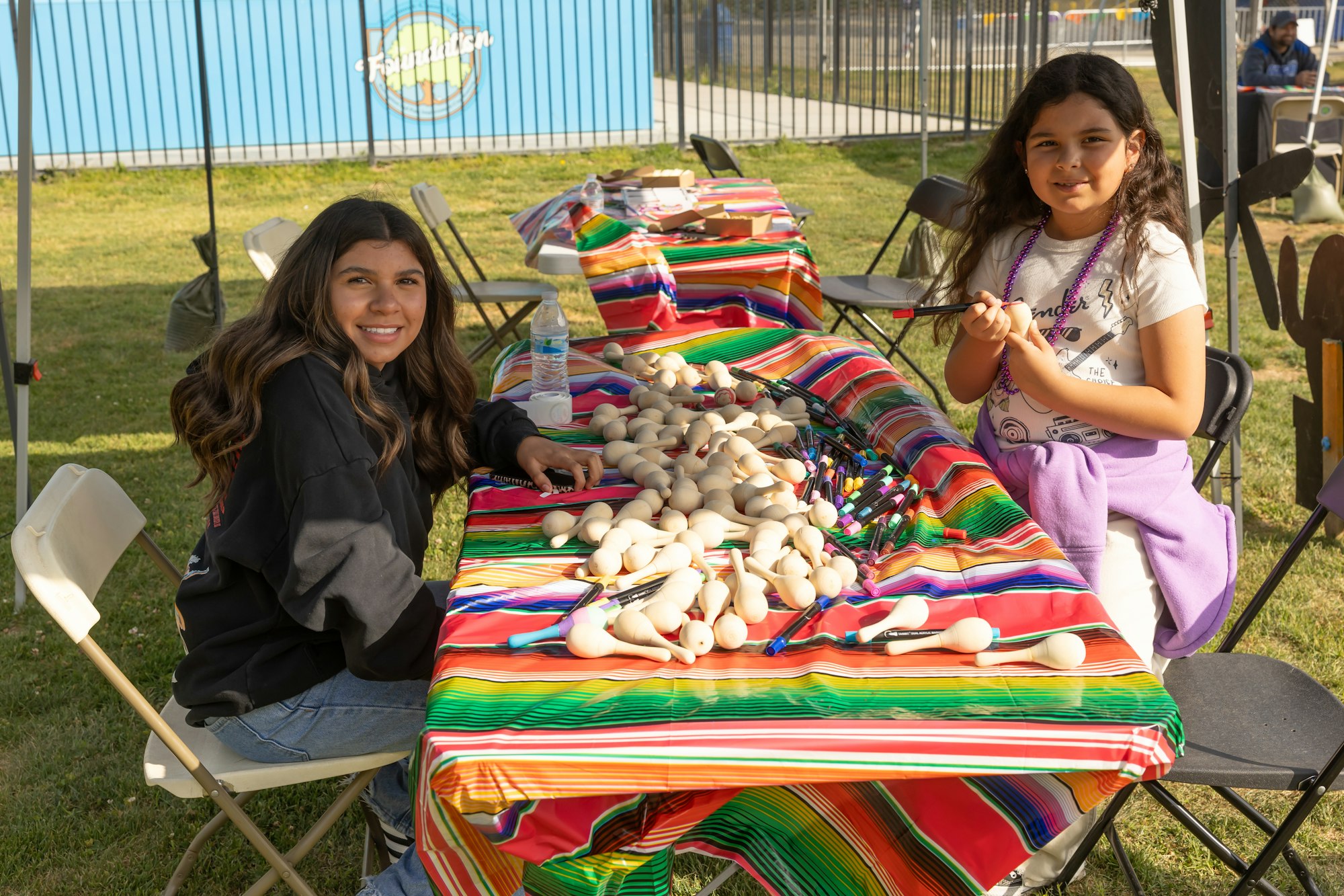 Two people at a colorful table with art supplies and wooden toys, outdoors.