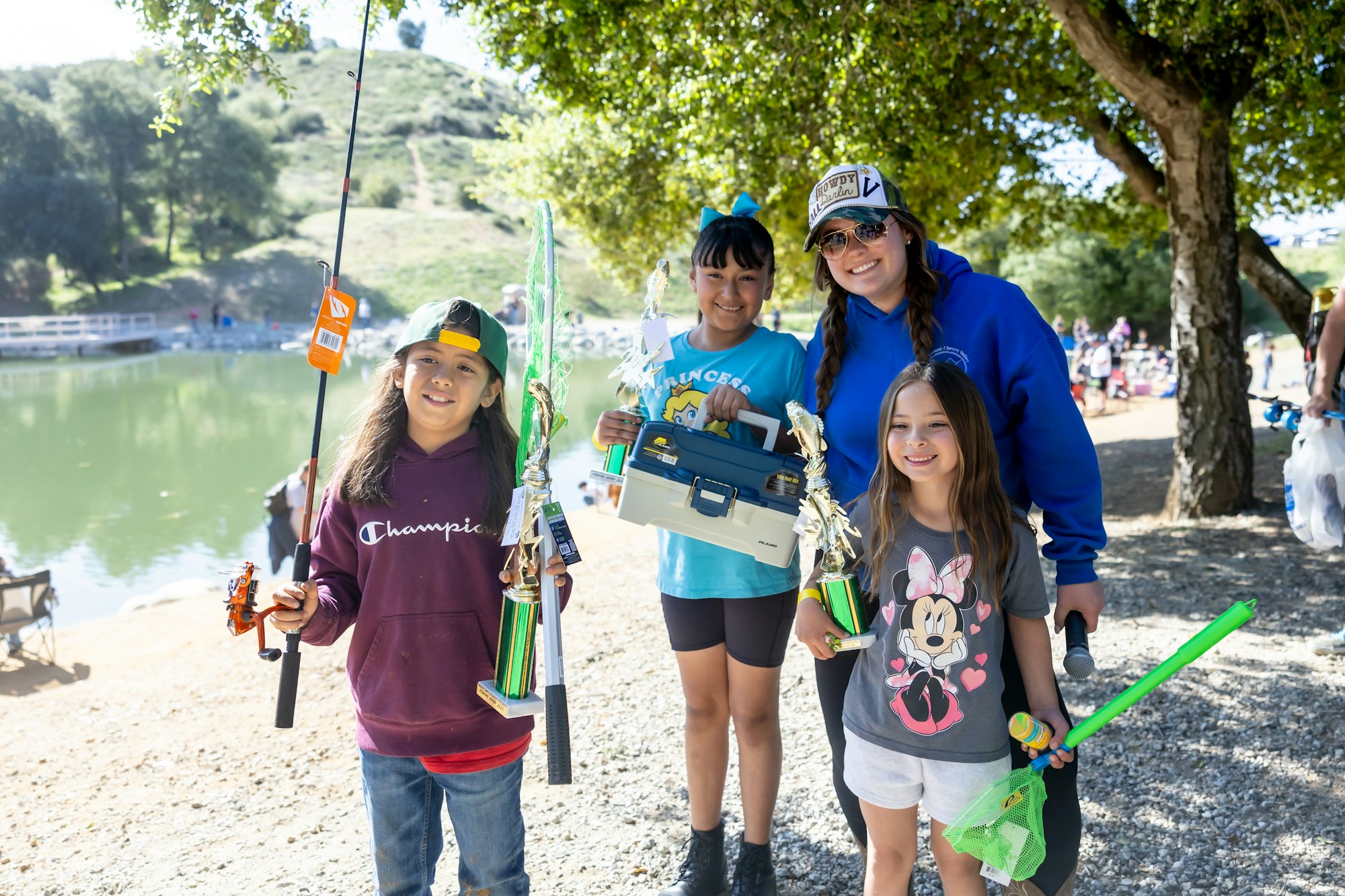 A group of people posing by a lake with fishing gear and trophies, smiling under a tree.