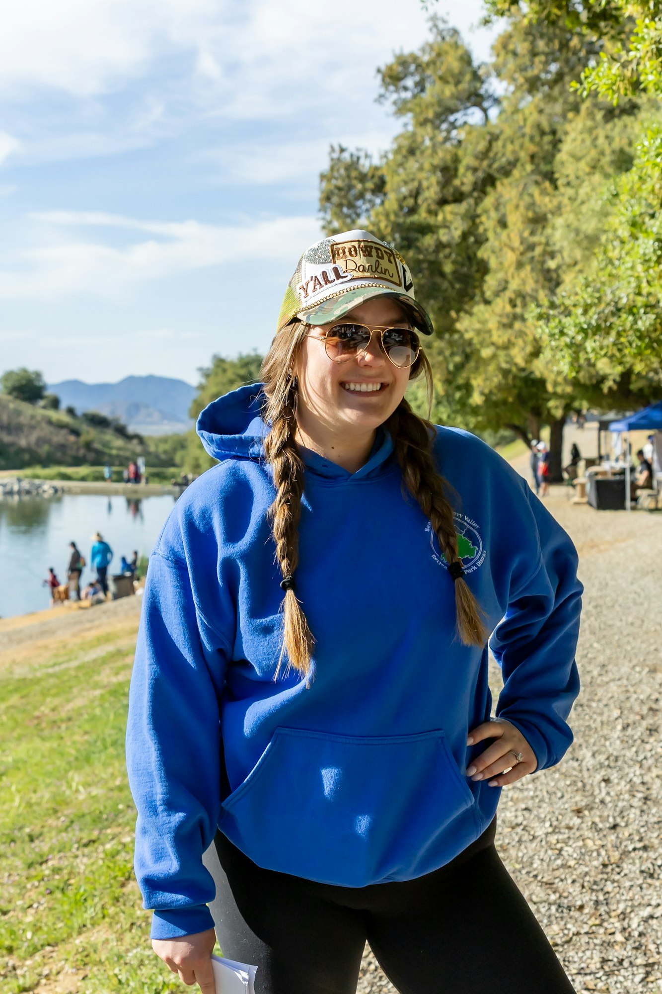 Person smiling by a lake, wearing sunglasses and a blue hoodie, with trees and people in the background.