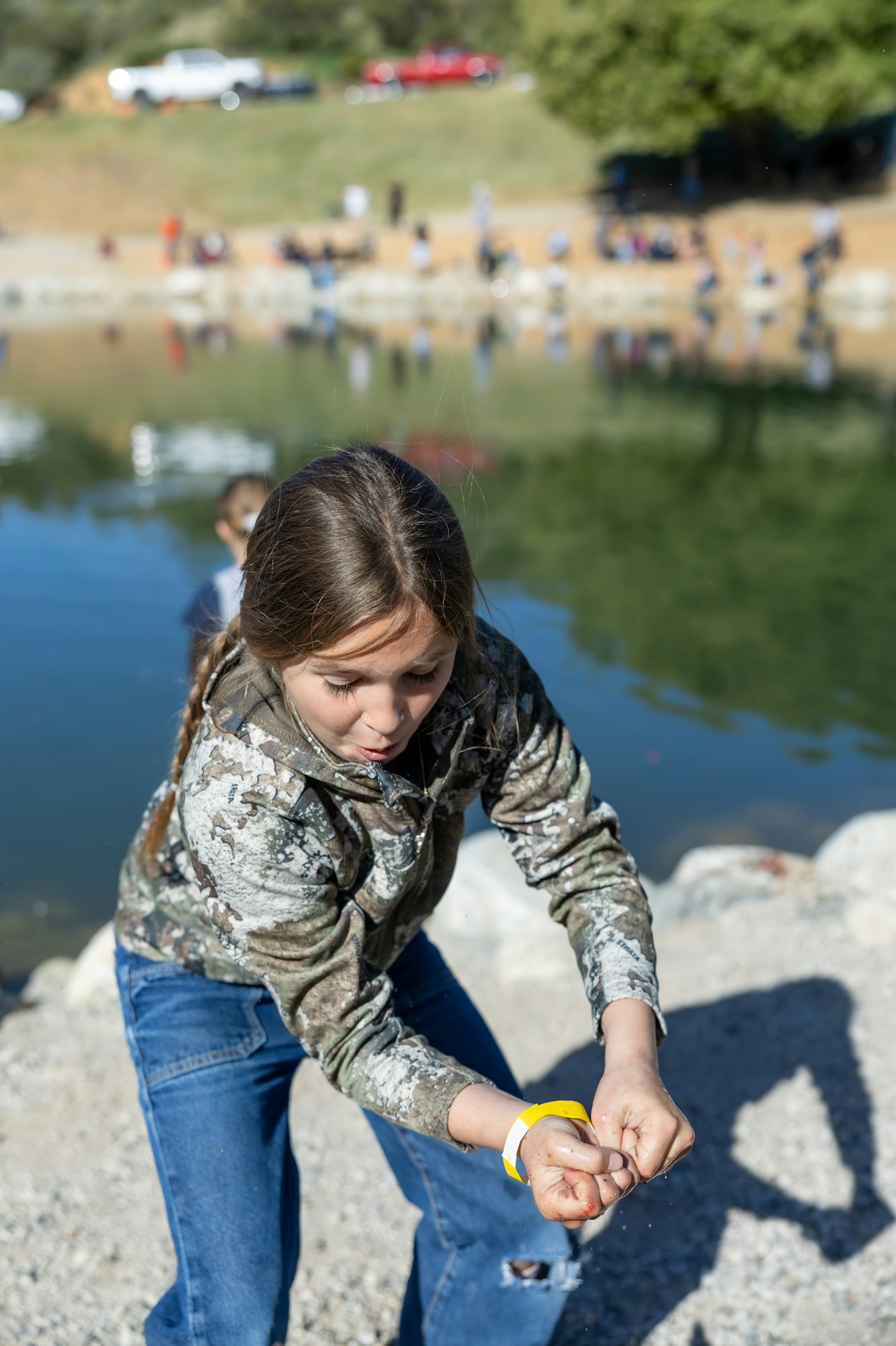 A person on a lakeside, dressed in a camo jacket and jeans, appears to be holding something while looking down.