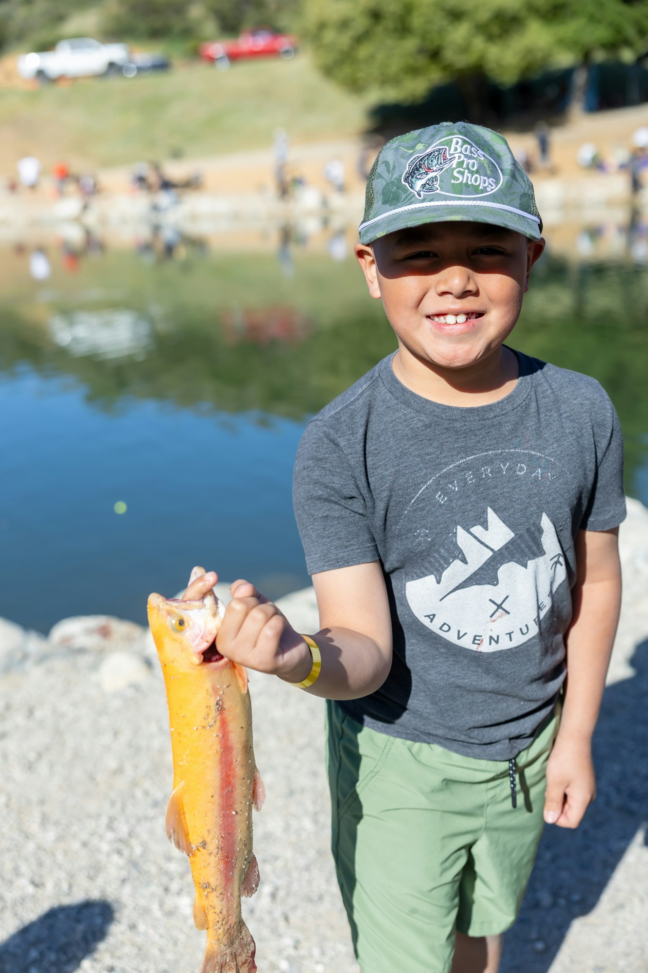 A smiling kid in a cap proudly holding a fish near a pond, with blurred people in the background.