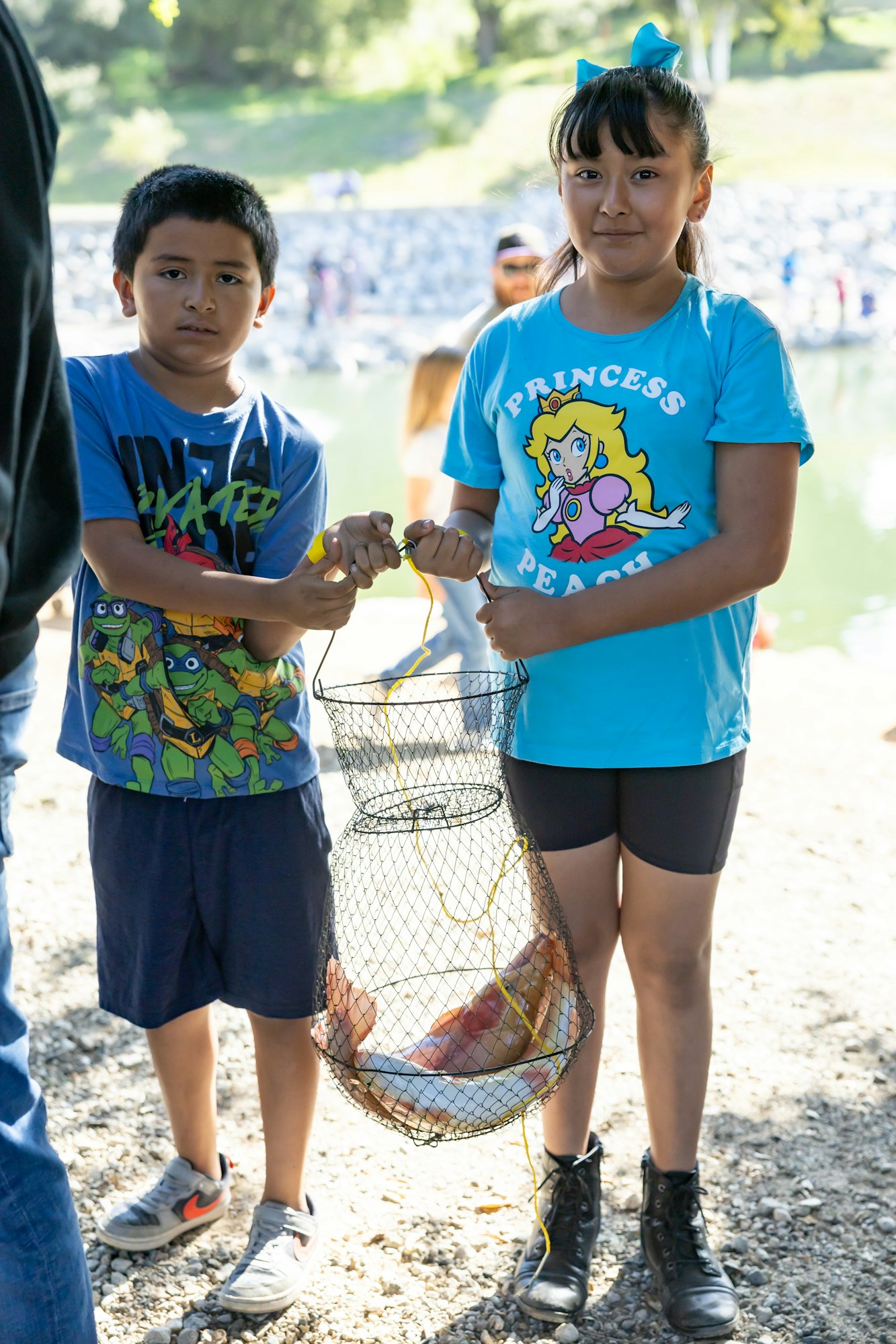 Two kids holding a basket with fish, wearing cartoon-themed shirts near a pond.