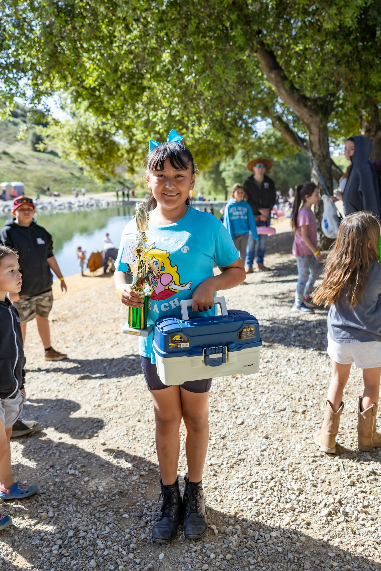 A child with a blue bow holds a trophy and a tackle box, standing outdoors with other people nearby.