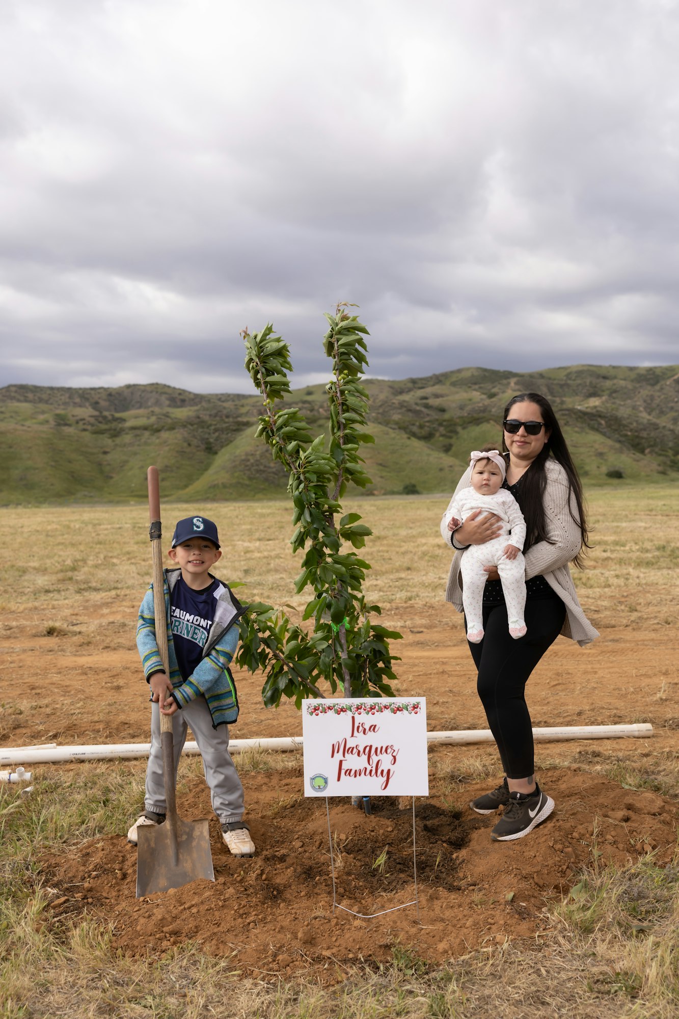 A woman holds a baby next to a young boy with a shovel, beside a small tree and a sign saying "The Marquez Family" in a field.