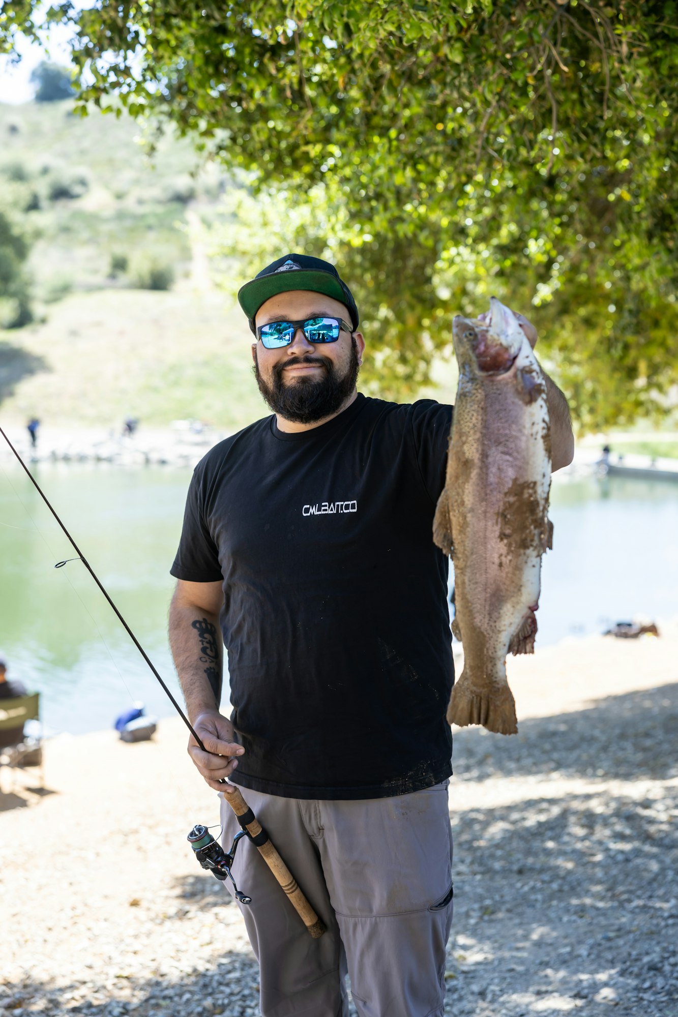 A person holds a large fish next to a body of water, wearing sunglasses and a cap, with a fishing rod in hand.