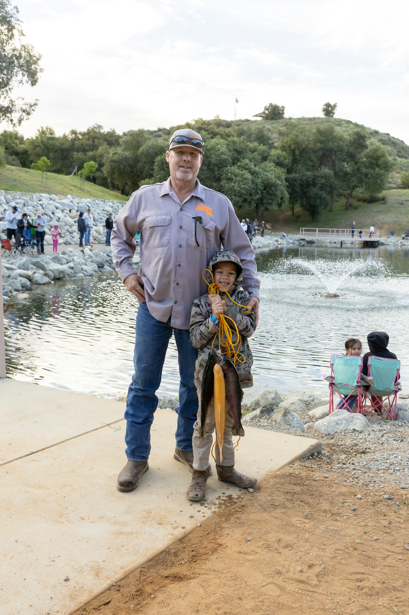 A man and child at a lake with captured fish, smiling. People in the background near water with fountains.