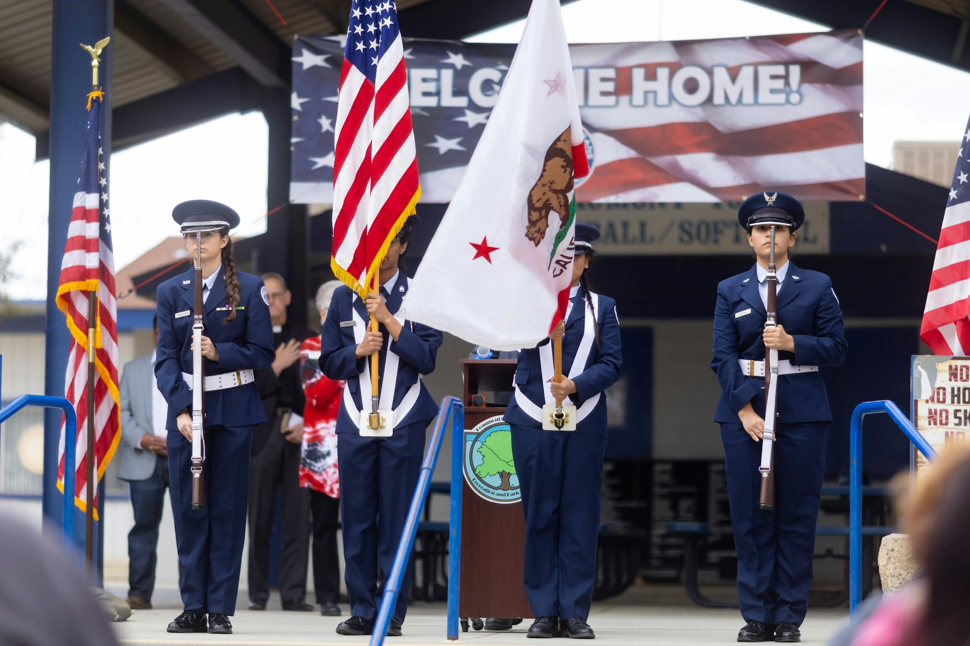 Uniformed color guard with U.S. and California flags, in front of a "Welcome Home" sign during a ceremony.