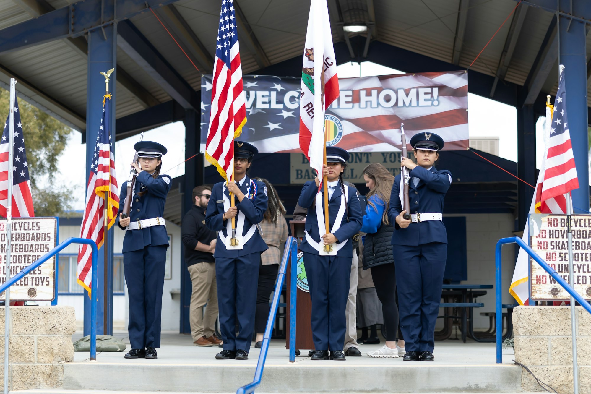 Color guard presenting flags at a ceremony with a "Welcome Home!" banner in the background.