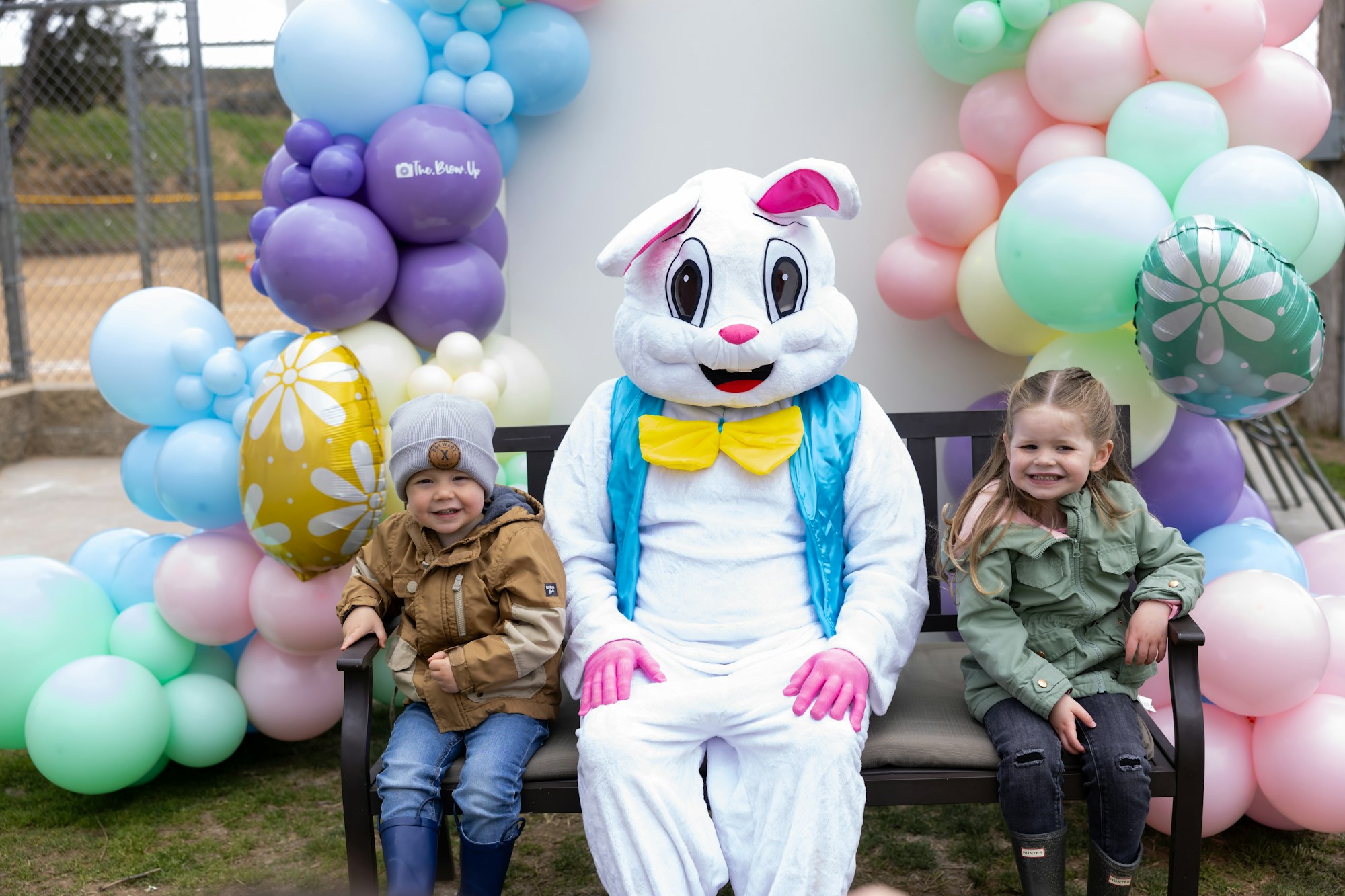 A person in an Easter Bunny costume sits on a bench with two smiling children, surrounded by colorful balloons.