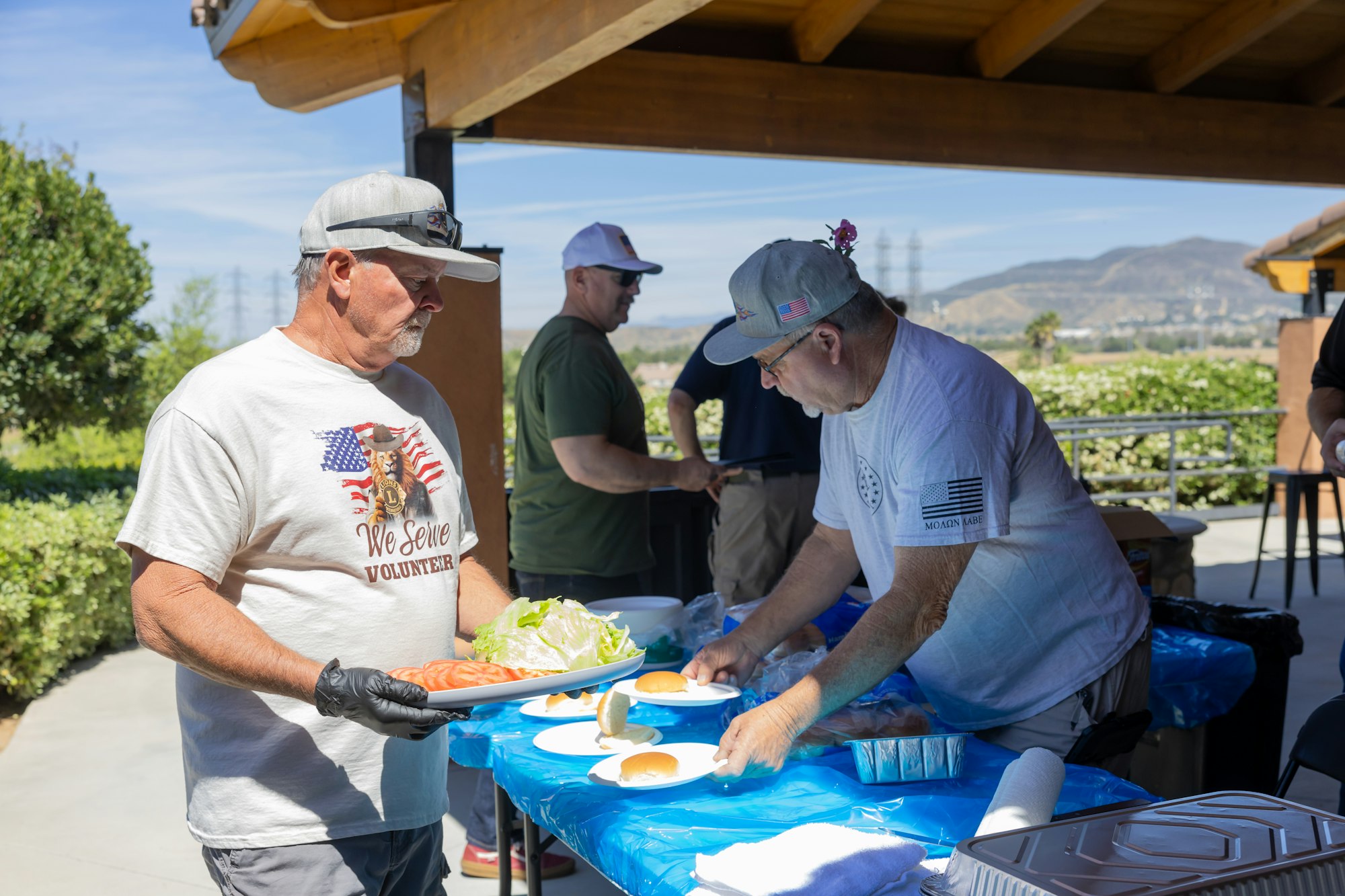 People are preparing food at a picnic or outdoor gathering, serving buns, lettuce, and tomatoes under a pavilion.