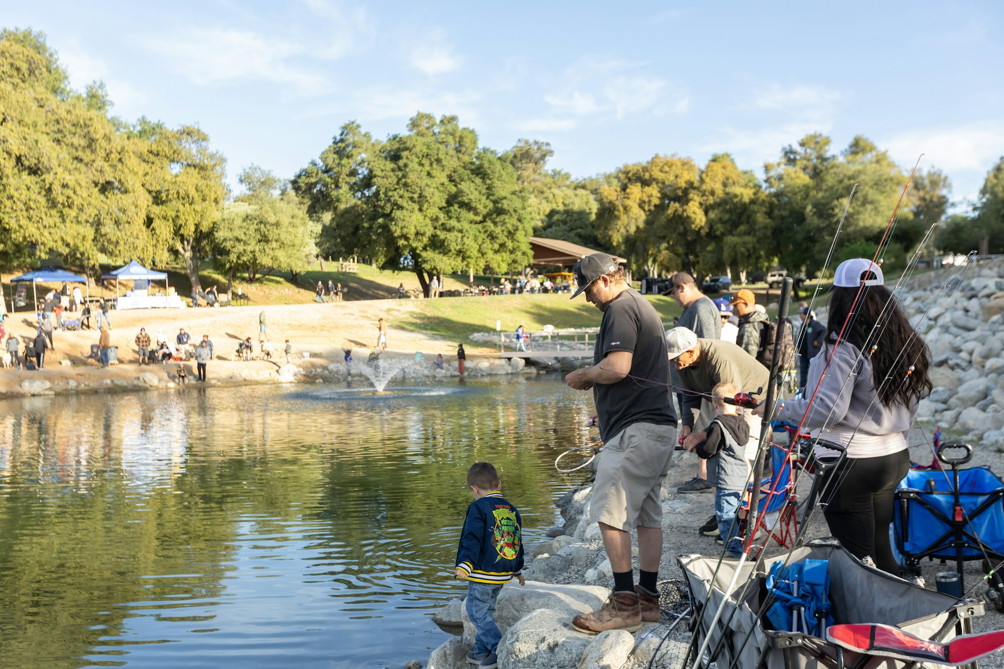 People fishing by a lake in a park, with trees, tents, and a fountain in the background.