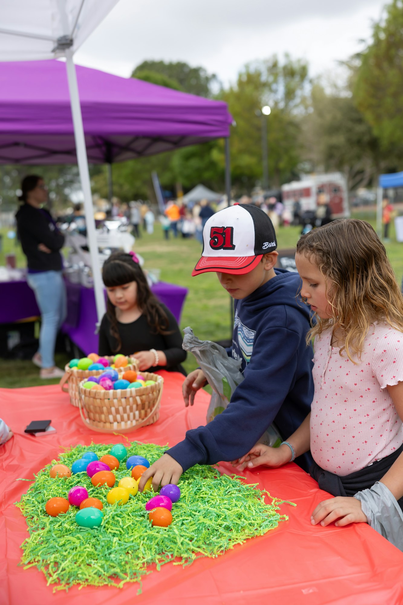 Children at a table with colorful eggs and baskets at an outdoor event under a purple tent.