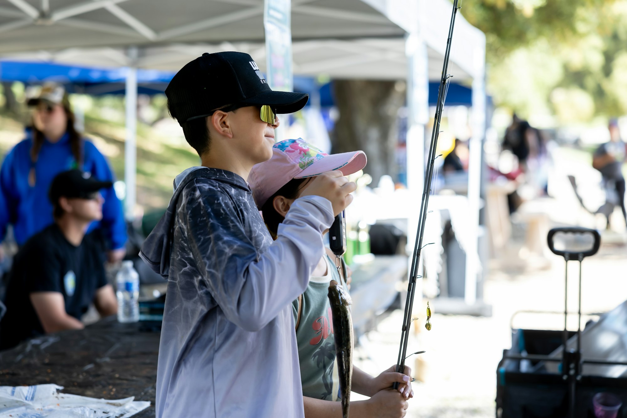Two kids wearing hats and sunglasses, holding a fishing rod and a fish, with people and tents in the background.