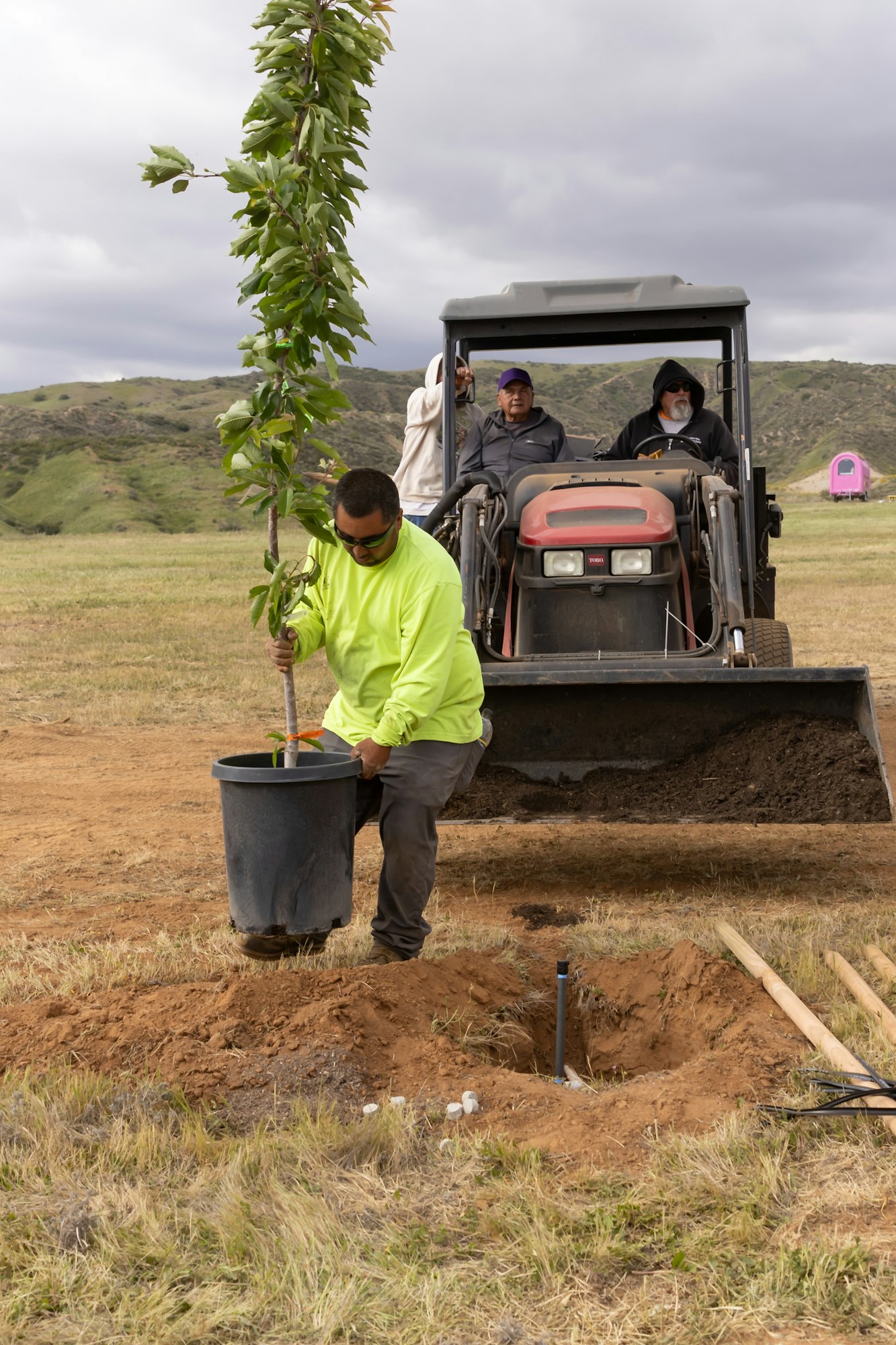 A person in a neon shirt planting a tree while others watch from a tractor in a grassy field.