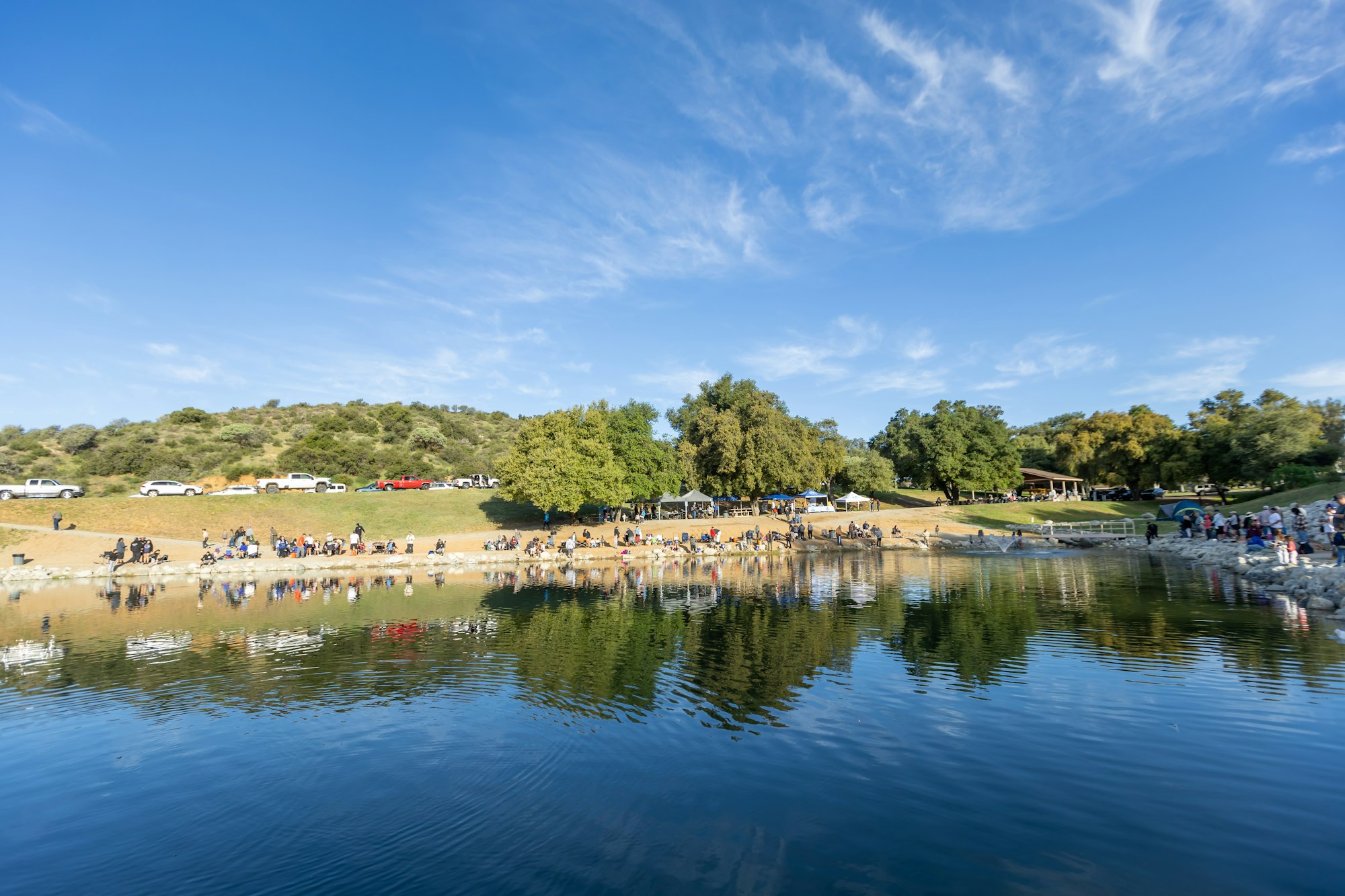 A scenic lake with people gathered along the shore, cars parked on a nearby road, and trees under a blue sky.