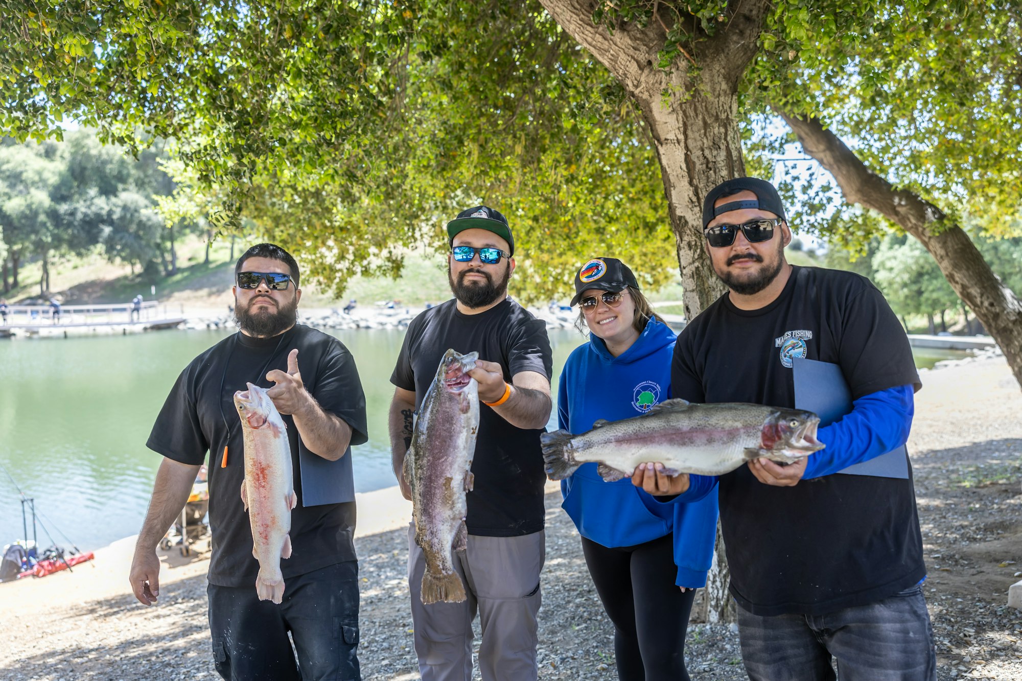 Four people holding fish by a lakeside, standing under a tree.